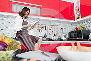 Asian cute middle-age woman in an apron stading using tablet computer connect to internet in kitchen with a smiling face and happy