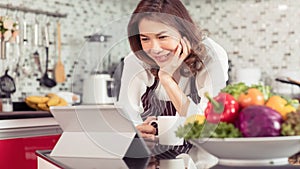Asian cute middle-age woman holding coffee cup and using tablet computer connect to internet in kitchen with a smiling face and