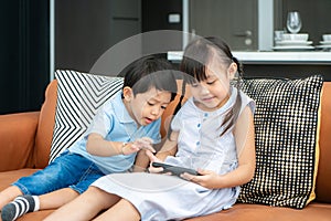 Asian cute sibling child using a smartphone and smiling for watching video or play game togerther while sitting on sofa in living