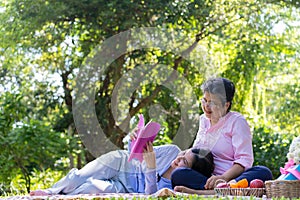 Asian daughter Sleeping on mother lap and reading book on picnic mat in the park. A happy senior woman talks with her daughter.