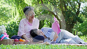 Asian daughter Sleeping on mother lap and reading book on picnic mat in the park. A happy senior woman talks with her daughter.