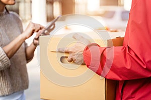 An Asian deliver man in red uniform handing parcel to a female costumer in front of the house. A postman and express delivery