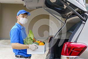 Asian delivery man wearing face mask and glove with groceries box of food, fruit, vegetable and drink standing near van in front