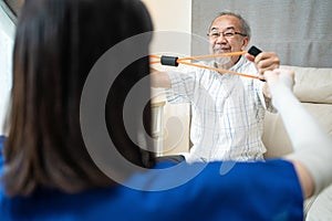 Asian Disabled senior older man sitting on sofa doing physiotherapist. Elderly handicapped man using resistance stretch band
