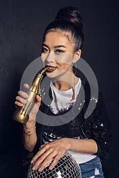 Asian disco girl with golden bottle and disco ball smiling and looking at camera isolated on grey