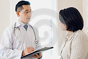 Asian doctor talking with middle aged woman in clinic asking symptoms for examination