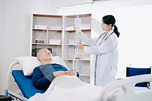 Asian doctor in white suit take notes while discussing and Asian elderly, man patient who lying on bed with receiving saline