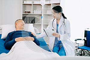 Asian doctor in white suit take notes while discussing and Asian elderly, man patient who lying on bed with receiving saline