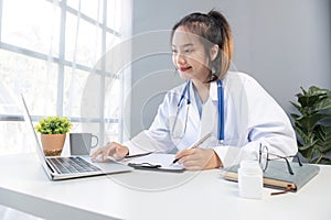 Asian doctor young beautiful woman smiling using working with a laptop computer and her writing something on paperwork