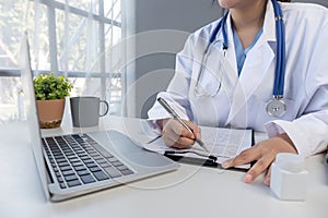 Asian doctor young beautiful woman smiling using working with a laptop computer and her writing something on paperwork