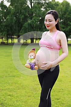 Asian Eastern Chinese happy pregnant woman in yoga dress hands hold her belly expecting baby to born plush toy bear beside excited