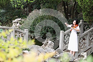 Asian Eastern Chinese young artist player woman carry play violin perform music park garden nature ancient building stand bridge