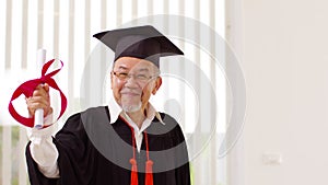 Asian elderly in cap and gown smile and celebration proud success graduate. Cheerful senior man holding certificated or diploma