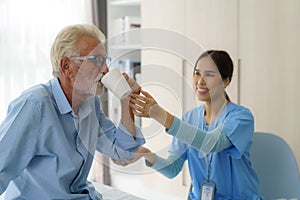 Asian elderly caregiver woman sitting on a hospital bed next to an older man helping drink a glass of water from the bedroom at