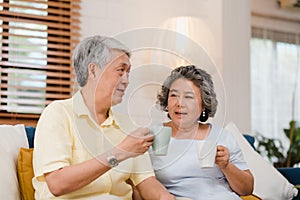 Asian elderly couple drinking warm coffee and talking together in living room at home, couple enjoy love moment while lying on