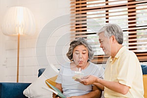 Asian elderly couple man holding cake celebrating wife`s birthday in living room at home. Japanese couple enjoy love moment