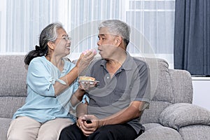 Asian elderly couple, are sitting on a sofa in home holding and eating donut