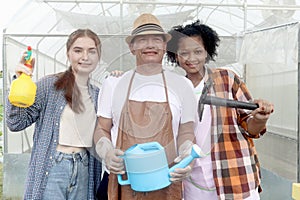 Asian elderly farmer hold watering can with two multiethnic teenager friend girls hold garden tools, standing at front of
