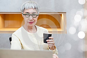 Asian elderly female gray hair wear spectacles holding coffee cup working on laptop computer at home office. Retired woman eating