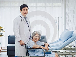 Asian elderly female patient dressed in light blue, smiling happily sits in wheelchair with kindly male doctor.