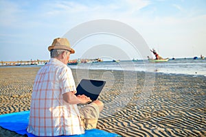 Asian elderly man sitting on the beach by the sea playing on his laptop computer