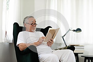 Asian Elderly man with white hairs sitting on sofa reading book ralax and cozy at home