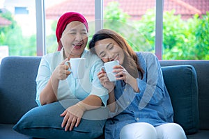 Asian elderly mother who recover from cancer and her daughter are sitting on a couch holding a cup of drink and talking to one