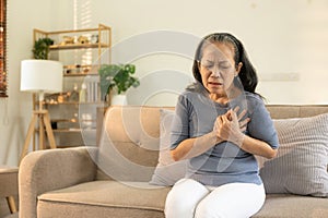 Asian elderly woman in gray casual clothes having left chest pain on sofa, senior mother