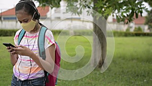 Asian elementary schoolgirl in protective face mask wearing headphones and standing at public park.
