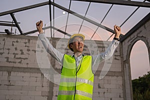 Asian engineer in uniform and helmet at construction site being happy and smiling for success.