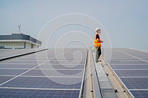 Asian engineer wearing protective vest and white hardhat standing on solar panels roof with blue sky background, Photovoltaic