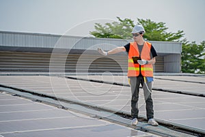 Asian engineer wearing protective vest and white hardhat standing testing heat while holding tablet on solar panels roof.