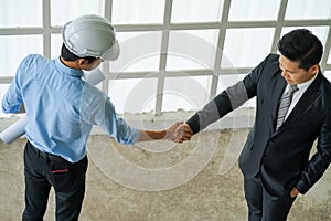 Asian engineer wearing white hard hat with blueprint meeting with business man for project