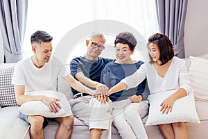 Asian family with adult children and senior parents putting hands together and sitting on sofa at home together. Family unity