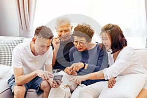 Asian family with adult children and senior parents using a mobile phone and relaxing on a sofa at home together.
