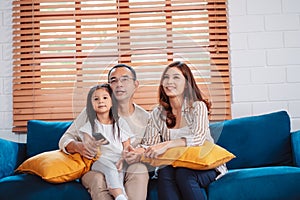 Asian family consisting of happy parents and daughter watching TV or movie together on sofa in living room at home. enjoy relaxing