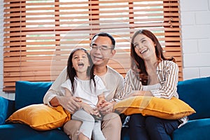 Asian family consisting of happy parents and daughter watching TV or movie together on sofa in living room at home. enjoy relaxing