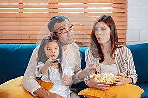 Asian Family consisting of parents, happy daughter watching TV or movie eating popcorn on sofa in living room at home. enjoy