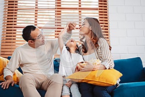 Asian Family consisting of parents, happy daughter watching TV or movie eating popcorn on sofa in living room at home. enjoy