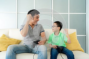 Asian family father and his son to playing video games with joysticks and arm raised to winning in game while sitting in sofa in