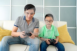 Asian family father and his son to playing video games with joysticks while sitting in sofa in living room at home, concept of