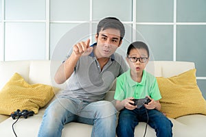 Asian family father teaching his son to playing video games with joysticks while sitting in sofa in living room at home, concept
