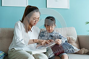 Asian family with mother and daughter reading book together while sitting on the sofa in the living room at home morning