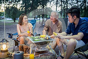 Asian family with senior drinking and having barbecue together. Cooking grilled bbq for dinner during camping on summer