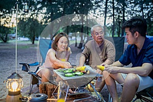 Asian family with senior drinking and having barbecue together. Cooking grilled bbq for dinner during camping on summer