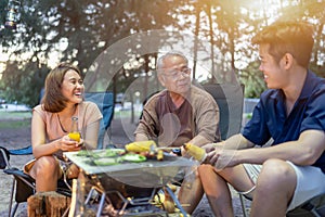 Asian family with senior drinking and having barbecue together. Cooking grilled bbq for dinner during camping on summer