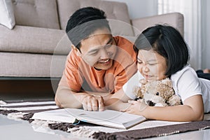 Asian father and daughter are reading book while lying on the carpet in the living room at her home. The cute little girl hugs the