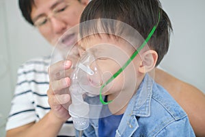Asian father helping his toddler son with inhalation therapy by the mask of inhaler. Sick little kid with respiratory problem with