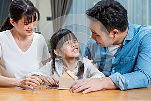 Asian Father and Mother teach young daughter to save money for future. Happy little girl enjoy put coins in piggy bank with