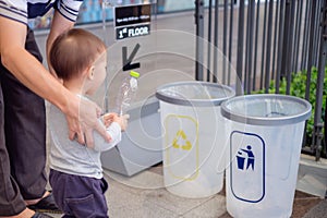 Asian father teach cute little 18 months, 1 year old toddler baby boy child throwing plastic bottle in recycling trash bin at publ